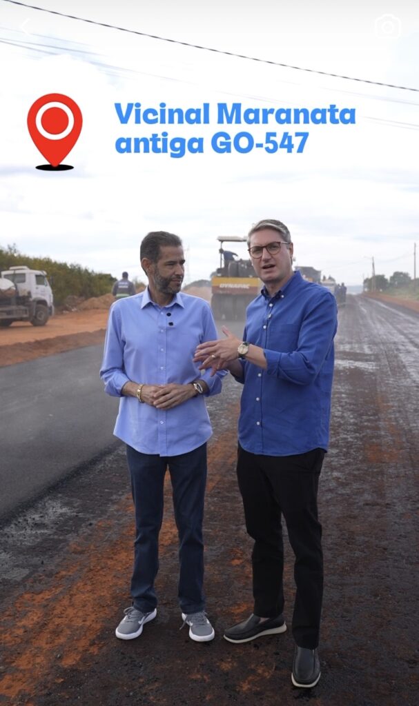 Fotografia mostrando o deputado estadual Anderson Teodoro acompanhando o andamento das obras na estrada de terra da Vicinal Maranata, antiga GO-547, em Águas Lindas de Goiás. Ao fundo, é possível ver o maquinário pesado trabalhando na pavimentação asfáltica. A imagem possui um selo com um ícone de localização vermelho e o texto do nome da via.