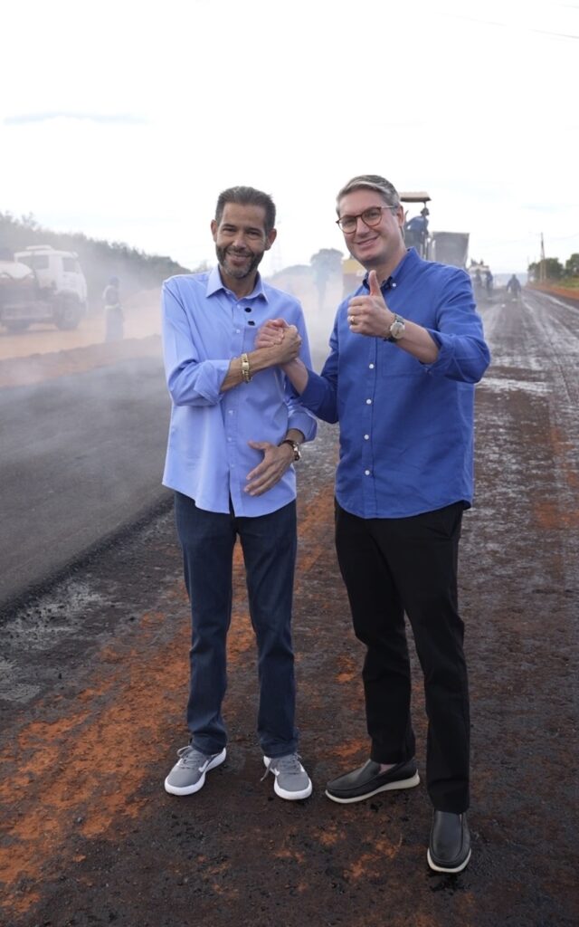 Fotografia do deputado estadual Anderson Teodoro e do prefeito de Águas Lindas de Goiás, Dr. Lucas Antonietti, acompanhando as obras de pavimentação. Os dois estão de pé na estrada de terra da Vicinal Maranata, cumprimentando-se, com o prefeito fazendo um sinal de positivo. Ao fundo, nota-se o maquinário pesado trabalhando na obra.
